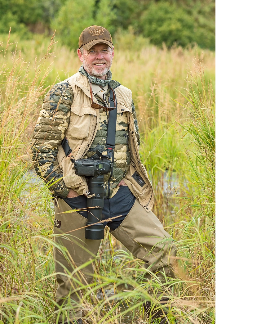 A person wearing a camouflage jacket, beige vest, and brown cap is standing in tall grass holding a camera with a large lens and smiling. Description generated by AI