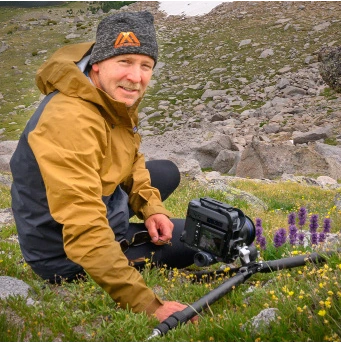 A person wearing a brown and black jacket and a gray beanie is sitting on grass and rocks in a mountainous area while adjusting a camera mounted on a tripod. Description generated by AI