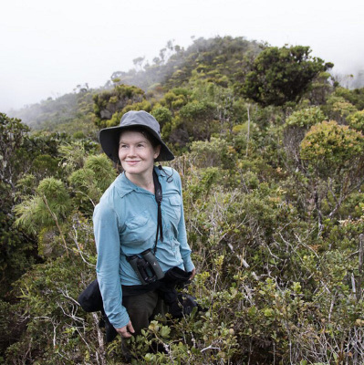 A person wearing a wide-brimmed hat and blue shirt stands smiling amidst dense green foliage on a misty hillside. Description generated by AI
