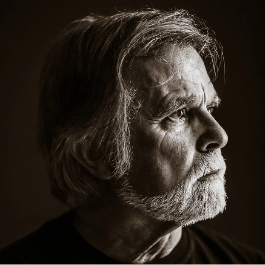 A senior person with medium-length hair and a beard is shown in a sepia-toned close-up profile looking thoughtfully into the distance. Description generated by AI