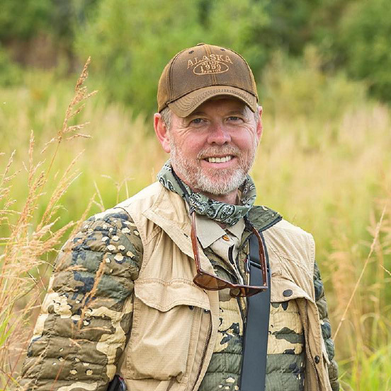 A person wearing a camouflage jacket, beige vest, a brown cap with “Alaska" written on it, and sunglasses hanging from their neck is smiling while standing in a grassy outdoor setting. Description generated by AI